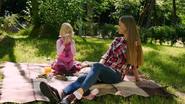Two sisters on a picnic outdoors. The little blond girl drinking juice and holding the thumb upwards. 4K