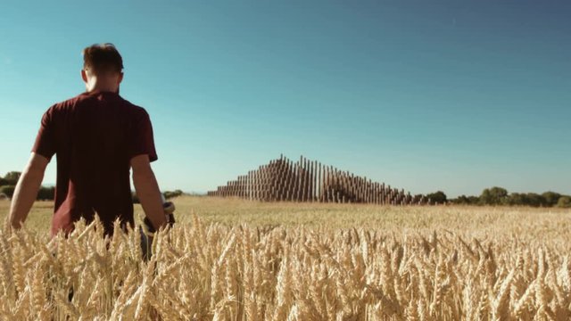 Adult walking through Beautiful wheat field towards modern pyramide holding skateboard with blue sky and epic sun light - shot on RED 4k Super Slow Motion