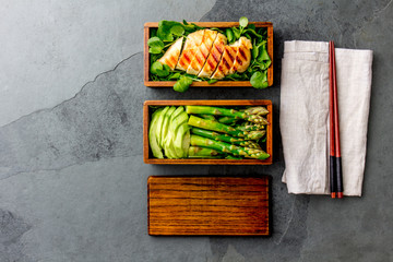 Healthy lunch in wooden japanese bento box. Balanced healthy food grilled chucken and avocado with asparagus and green salad. Top view, slate gray background