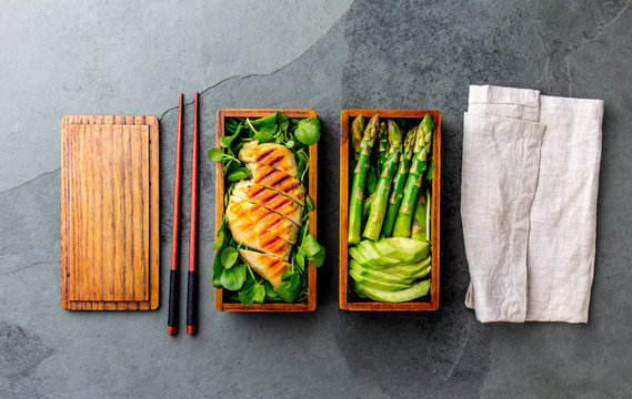 Healthy lunch in wooden japanese bento box. Balanced healthy food grilled chucken and avocado with asparagus and green salad. Top view, slate gray background
