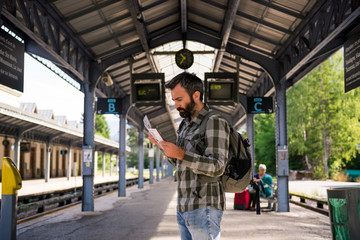 Young adult men commuter waiting for train in mountain train station reading timetable in sunny summer day.
