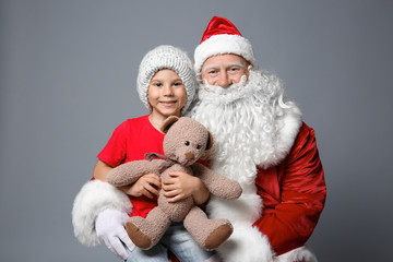 Cute little boy with teddy bear sitting on Santa's lap against color background
