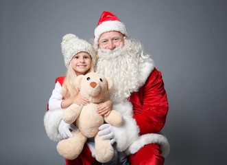 Cute little girl with teddy bear sitting on Santa's lap against color background
