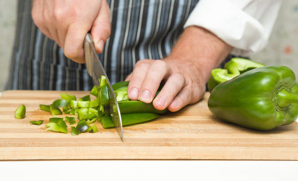Chef's hands with knife cutting green paprika on the wooden board on the white table in the kitchen. Cooking concept. 