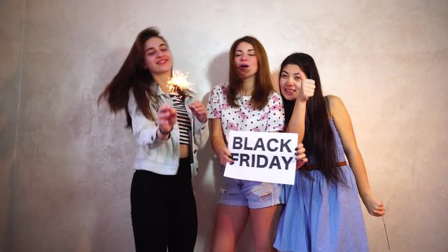 Lovely girls with smiles on their faces look at camera and pose, hold sign in their hands denoting with Black Friday texts, discounts and purchases, stand in room against background of light wall