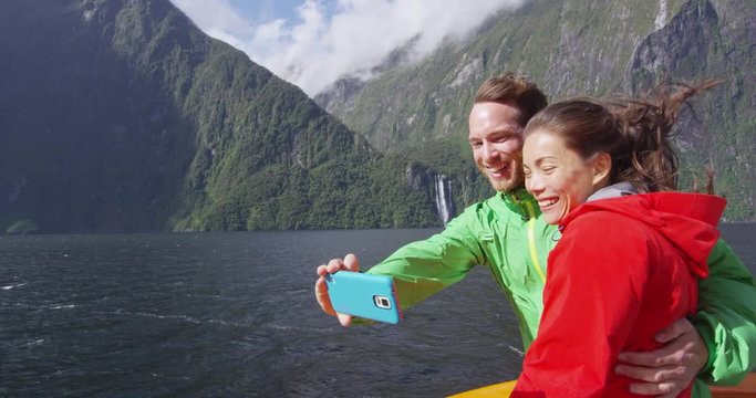 Happy couple taking selfie using smart phone on cruise ship, Milford Sound, Fiordland National Park, New Zealand. Multicultural couple smilng having fun laughing on travel vacation. SLOW MOTION.