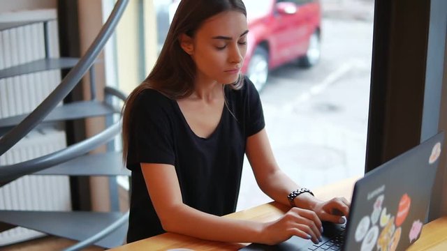 Young woman sitting in the coffee shop by the window with her laptop and thinking what to write or how to answer. Then she has an idea and starts to type