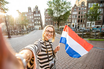 Obraz premium Young smiling woman tourist making selfie photo standing with dutch flag on the beautiful buildings background in Amsterdam city