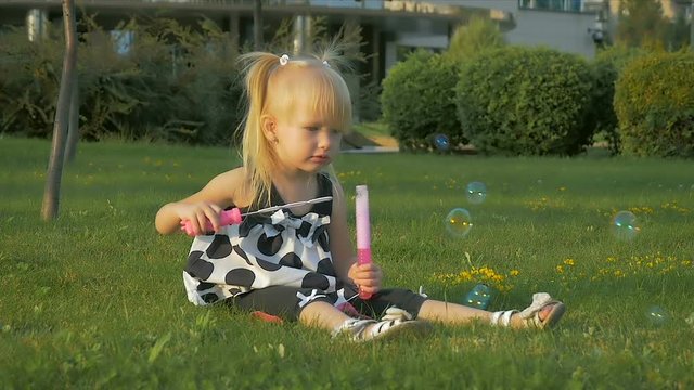 SLOW MOTION: little girl of two-years is sitting on the grass and blowing soap-bubble in the city park.