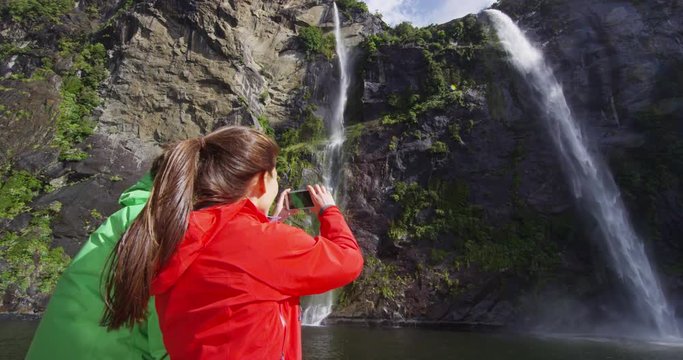 Cruise ship tourists taking photos using phone on boat tour in Milford Sound, Fiordland National Park, New Zealand. Couple sightseeing travel sailing by fairy falls waterfall on New Zealand.