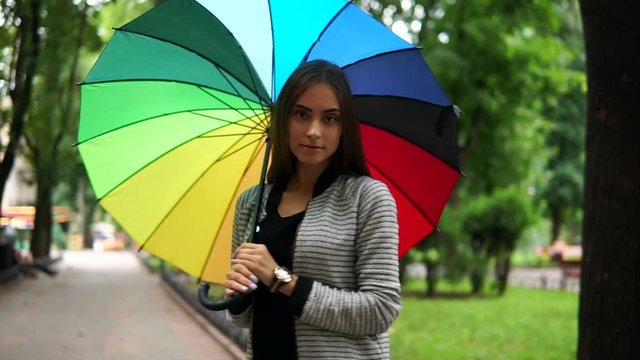Portrait of a young attractive brunette woman spinning her colorful umbrella in a rainy day in the city park and looking in the camera