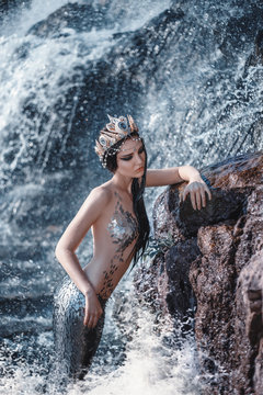 Portrait Fantasy woman beauty face real mermaid is resting on ocean shore. Silver fish tail, body covered with scales. Creative photo splash weves water sea stone. Creative costume crown from shells