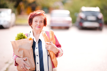 Happy senior woman 70-80 year old holding package of food walking on street outdoors. Looking at camera. Healthy eating.
