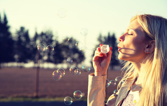 Gorgeous young blond girl blowing soap bubbles in sunlit park.
