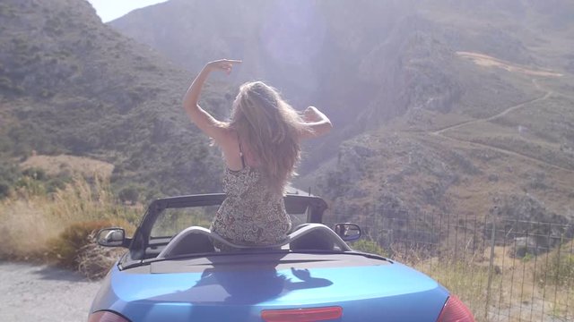 Rear view of relaxing woman with her hands up sitting in blue cabriolet car.Vacation, holiday, journey concept. Slow motion shot of young beautiful girl with long blonde hair.
