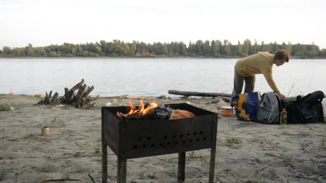 Man supports the fire in the grill. Family is resting on the river bank