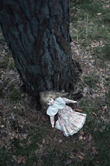 Spooky doll girl on forest ground against tree trunk.