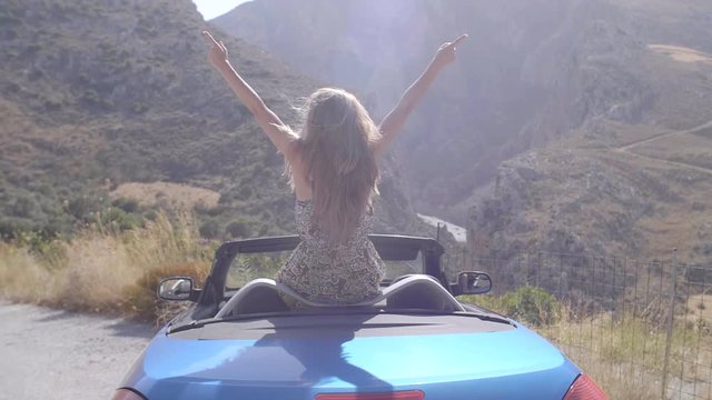 Rear view of relaxing woman with her hands up sitting in blue cabriolet car.Vacation, holiday, journey concept. Slow motion shot of young beautiful girl with long blonde hair.