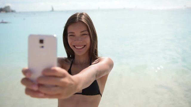Beach bikini girl taking selfie with smartphone smiling happy. Mixed race Asian Caucasian woman self portrait photograph with mobile cell smart phone on summer travel holidays vacation.
