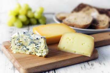 various cheese on a wooden board, bread and grapes blurred in the bright rustic background