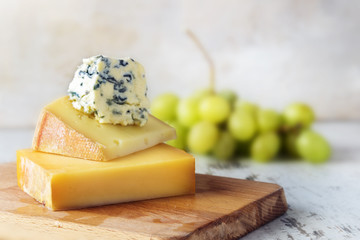 stack of various cheese on a wooden board, green grapes blurred in the bright background, copy space