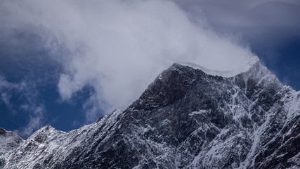 Panoramic view of a mountain peak covered with a snow on the top. Blowing snow and clouds blending above peak. Summer in the Pennine Alps, Valle d'Aosta, Italy, Europe.