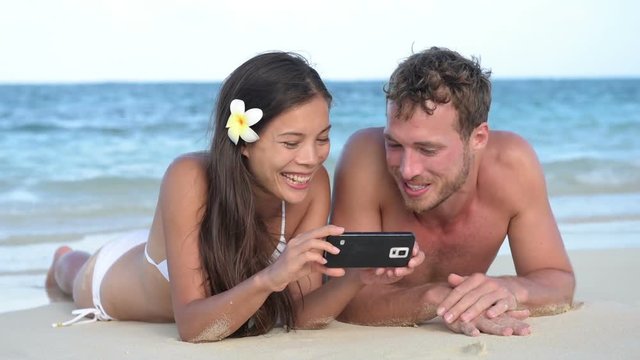 Beach holiday couple using smartphone having fun laughing looking at mobile cell smart phone screen lying down in sand. Young multicultural Asian woman and Caucasian man.