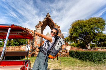 Fototapeta premium Young asian female traveler with backpack traveling with standing on taxi or Tuk Tuk and happy fun with old temple (Wat Mahathat) background, Ayutthaya Province, Thailand