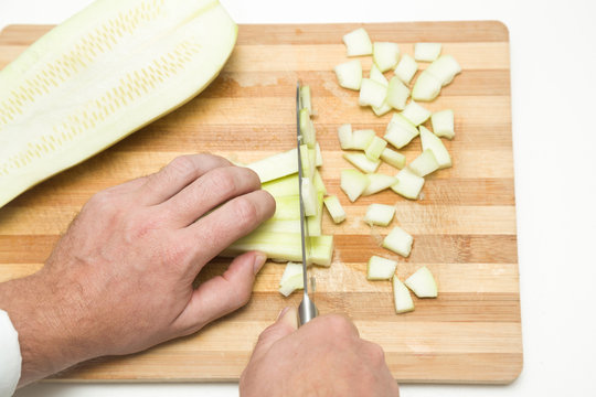 Chef's hands with knife cutting zucchini on the wooden board on the white table in the kitchen. Cooking concept.