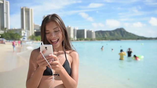 Earphones using woman with smartphone having fun on beach listening to music. Girl in bikini using mobile cell smart phone happy. Beautiful Asian Caucasian female model on Waikiki, Oahu, Hawaii, USA
