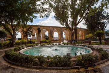 Sparkling fountain under peaceful trees and limestone arches on the background, Upper Barrakka Gardens park, Capital city of Malta, Valletta.