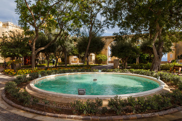 Sparkling fountain under peaceful trees and limestone arches on the background, Upper Barrakka Gardens park, Capital city of Malta, Valletta.