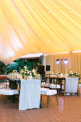 illumination, celebration, happiness concept. restaurant under large yellow like sunset tent prepared for banquet, all the tables are served with dishes and flowers, and spotlights are turned on