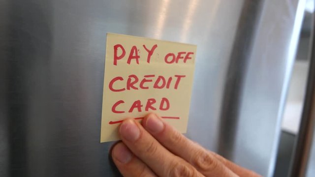 Putting a PAY OFF CREDIT CARD sticky note reminder on a fridge. Closeup on the hand and paper.