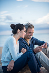 Obraz premium A couple sitting at the beach, looking at photos on a phone