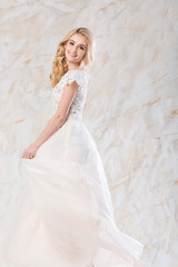 fashionable wedding dress, beautiful blonde model, bride hairstyle and makeup concept - happy young woman in long luxury white gown indoors on light background, stunning female posing with a smile