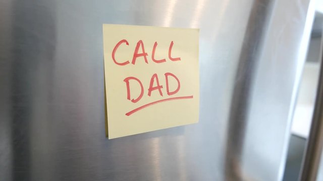 Putting a Call Dad sticky note reminder on a fridge. Closeup on the hand and paper.