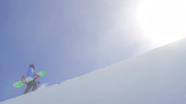 SLOW MOTION: Young pro snowboarder walking uphill in the halfpipe snow park carrying his board on a beautiful sunny day in snowy winter