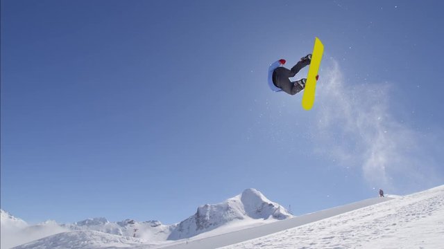 SLOW MOTION: Young pro snowboarder riding the half pipe in big mountain snow park, jumping out of the halfpipe wall and over the sun, performing tricks and rotations with grabs in sunny winter 