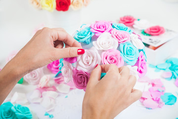 Young woman makes a bouquet of artificial flowers - roses from foam blue, pink, white, color in a white pot for decorating interiors, interiors and holidays, on the table lies a gun for glue, petals