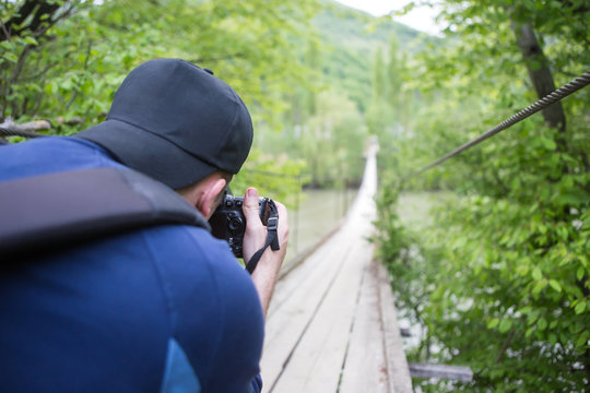 Traveler with backpack taking a photo of wooden suspension bridge over mountain river with professional camera 
