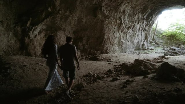 A guy with a girl holding hands exits the cave. A loving couple strolls along the cave.