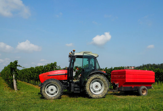 Red tractor with the door open and a red trailer in front of vineyards under blue sky with clouds