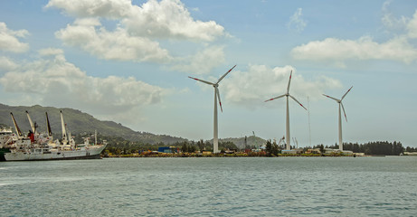 A waterfront with three wind turbines and a fishing ship near the green hills, Seychelles