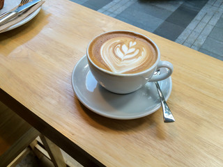 Cup of hot latte coffee with latte art on wooden table inside restaurant in morning light. Morning coffee illustrating modern lifestyle / diet