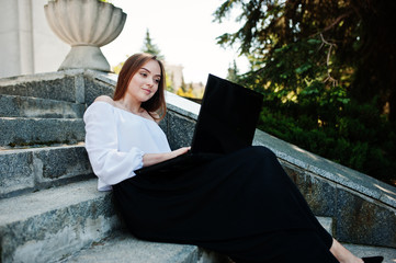 Good-looking young woman in white blouse, wide black pants and black classic high heels sitting on stairs and working on her laptop.