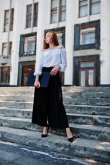 Portrait of a flawless young woman in white blouse, wide black pants and classic high heels posing on the stairs with black laptop in her hand with a huge white building on a background.