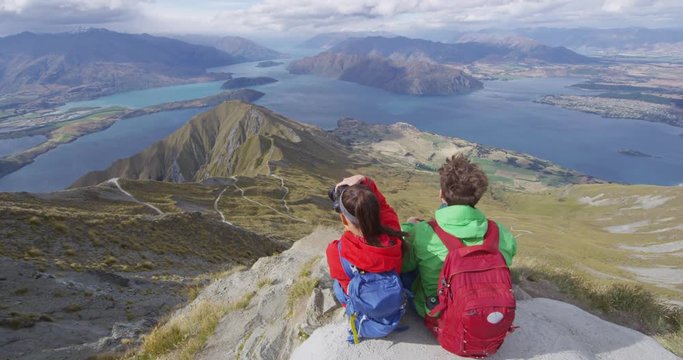 Hikers looking at view on mountain top summit on hiking travel vacation - couple taking slr camera photo. Wanderlust adventure people relaxing. Summit of famous hike to Roys Peak, New Zealand.