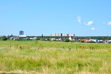 Fototapeta na wymiar Green uncultivated field with one-story houses and high-rise buildings in the background