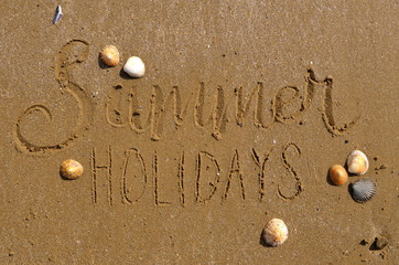 SUMMER HOLIDAYS hand lettered in sand on beach with shells